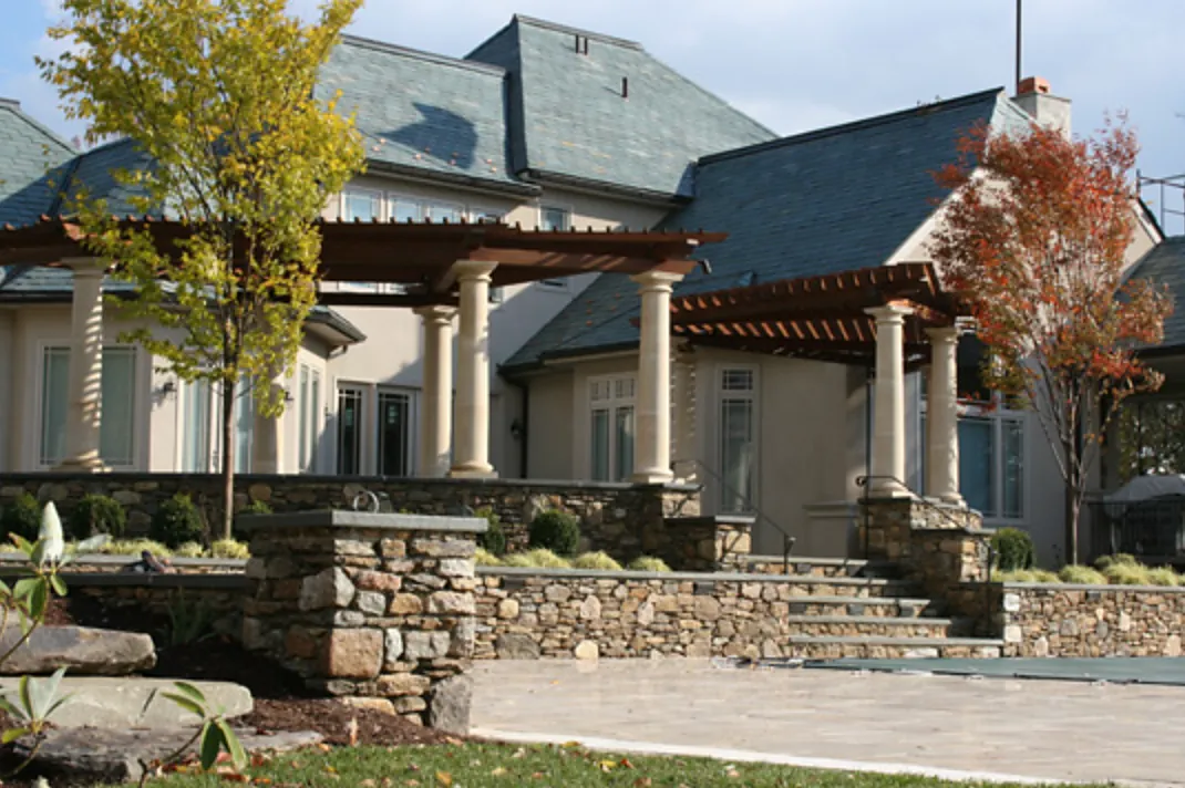 Fieldstone terrace walls, piers and steps below twin pergolas on a slate-roofed home